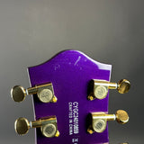 Close-up of the headstock of a purple electric guitar with gold tuning pegs and white printed model text on a gray background.