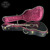 An acoustic guitar with a black body and gold hardware, displayed inside an open black hard case lined with shiny magenta velvet. The case is open to show the interior, and the guitar is positioned horizontally underneath.