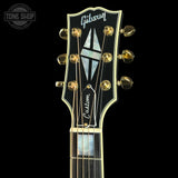 Close-up of the headstock of a Gibson Custom electric guitar against a solid black background. The headstock is black with gold hardware, a mother-of-pearl inlay, and the Gibson logo at the top.