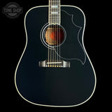 A close-up of a black acoustic guitar body with white binding, a natural wood soundboard visible through the round soundhole, and a white pickguard, shown against a black background with a Tone Shop watermark in the top left.