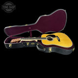 An acoustic guitar with a natural wood top and black body, displayed inside an open black hard case with a burgundy velvet interior, set against a solid black background.