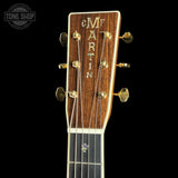 Close-up of a Martin acoustic guitar headstock with a dark brown wood grain finish, gold tuning pegs, and gold lettering, shown against a solid black background.