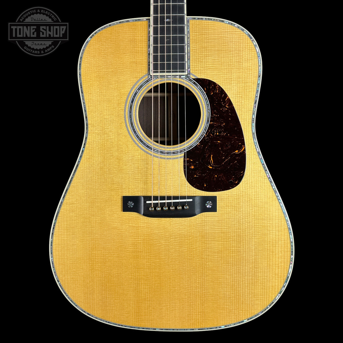 A close-up studio shot of an acoustic guitar body against a solid black background, featuring a natural wood finish, a dark tortoiseshell pickguard, and a silver-toned metal bridge.