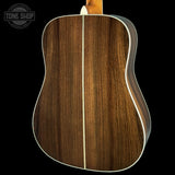 Close-up of the back of an acoustic guitar showing a rich dark brown wood grain finish with a light cream binding, set against a solid black background.