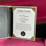 A Gibson Custom Certificate of Authenticity insert shown inside an open presentation folder, placed on a bright pink satin-like fabric background. The certificate identifies a Gibson Custom Dealer Select instrument and includes a Gibson logo seal.