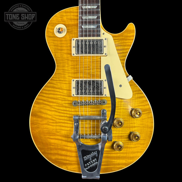 A close-up of a Gibson Les Paul Custom electric guitar featuring a vibrant yellow-orange flamed maple top, cream pickguard, and a silver-toned Bigsby tailpiece, set against a solid black background.