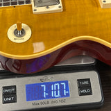 Close-up of a yellow-orange electric guitar body resting on a digital scale displaying 7.107 oz.