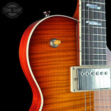 Close-up of a double-cutaway electric guitar body with a flamed maple wood finish in a vibrant orange-brown sunburst, featuring a white binding, a gold toggle switch, and a silver humbucker pickup against a black background.