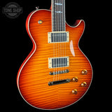 A close-up of a Les Paul style electric guitar body in a vibrant orange-to-brown sunburst finish, featuring gold hardware and a black pickguard, set against a solid black background.