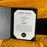 A Gibson Custom Certificate of Authenticity insert shown inside an open black folder on a textured mustard-yellow fabric background. The insert includes handwritten details for the LP Axxess Custom and a Gibson Select logo at the bottom.