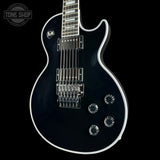 A black electric guitar with a glossy finish, chrome hardware, and white binding, shown against a solid black background with a Tone Shop watermark in the top left corner.