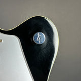 Close-up of a glossy black guitar body with a silver metal button featuring a lightning bolt symbol, set against a neutral gray background.