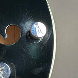 Close-up of a glossy black guitar body with a chrome tuning peg and a small circular logo emblem on the peg head, alongside a cutout headstock opening.