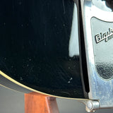 Close-up of a black electric guitar body with visible wear, scratches, and a silver Bighead Zender headstock edge.