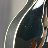 Close-up of a glossy black mandolin body with a gold-toned f-hole opening and a light-colored edge trim, showing some surface scratches.