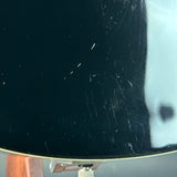 Close-up of a glossy black guitar body showing visible scratches and wear, with a small portion of a wooden headstock and metal hardware visible at the bottom.