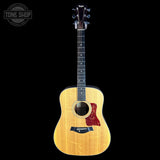 A Taylor acoustic guitar with a natural wood finish, black neck, and red pickguard, shown against a solid black background with a Tone Shop watermark in the top left corner.