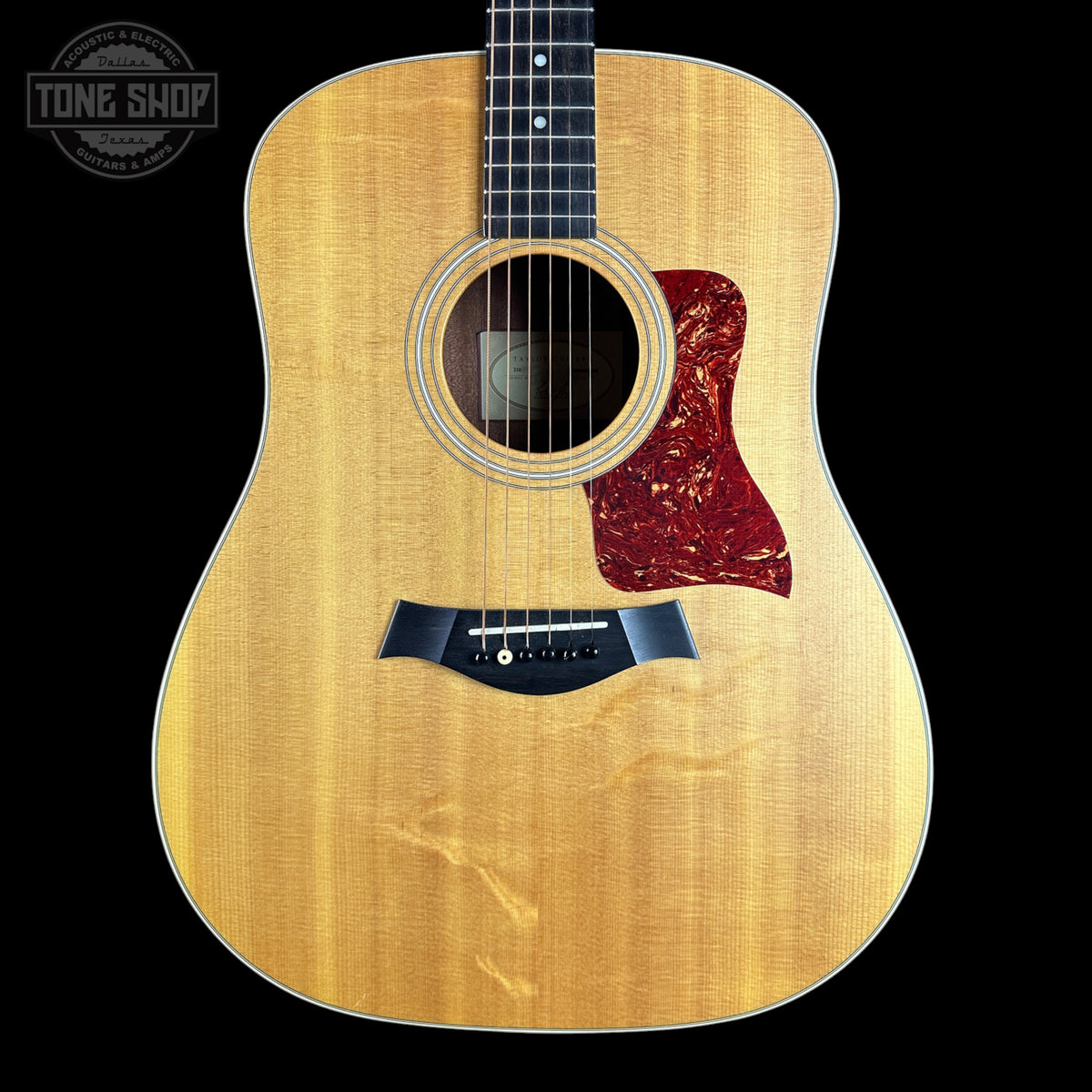 Taylor acoustic guitar with a natural flamed wood top, black pickguard, and red tortoiseshell-style pickguard, shown against a solid black background with a Tone Shop watermark in the upper left.