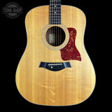 Taylor acoustic guitar with a natural flamed wood top, black pickguard, and red tortoiseshell-style pickguard, shown against a solid black background with a Tone Shop watermark in the upper left.
