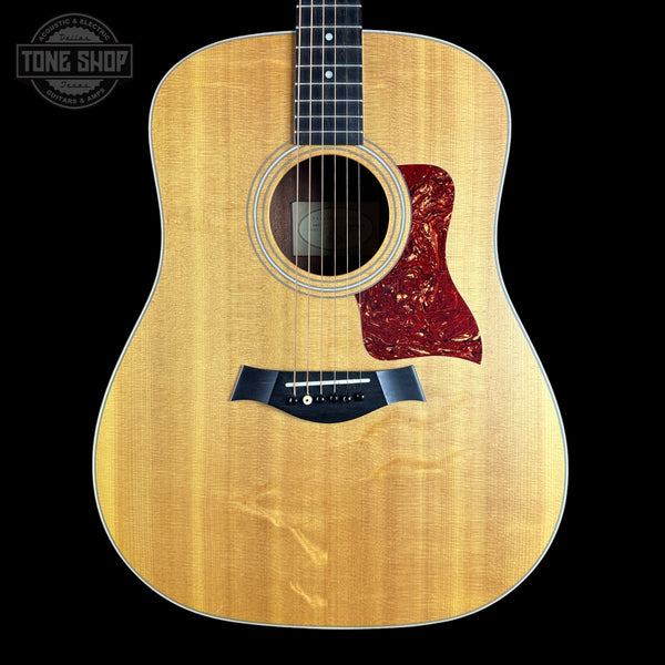Taylor acoustic guitar with a natural flamed wood top, black pickguard, and red tortoiseshell-style pickguard, shown against a solid black background with a Tone Shop watermark in the upper left.