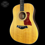 A Taylor acoustic guitar with a natural flamed top wood finish, a red maple pickguard, and a black bridge, shown against a solid black background with a Tone Shop watermark in the top left corner.
