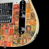 Close-up of an electric guitar body featuring a collage of vintage postage stamps in red, orange, green, and brown tones, with black hardware and control knobs on a white guitar body.