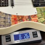 Close-up of a white electric guitar body with a vintage postage stamp collage print on the lower section, resting on a digital scale showing 8 lbs 26 oz.