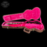 An open brown guitar case with bright hot pink plush lining, containing a dark electric guitar, shown on a black background with a Tone Shop watermark in the top left corner.