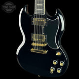 A black electric guitar with gold hardware and a white binding, shown from a high angle against a black background. A Tone Shop watermark is visible in the top-left corner.
