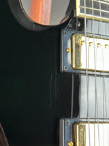 Close-up of a black electric guitar body showing the edge of the pickguard, gold humbucker pickups, metal strings, and part of the fretboard.