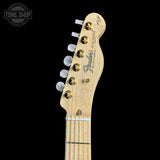 Close-up of the headstock of a Fender Telecaster electric guitar, showing the natural maple wood neck, gold hardware, and white tuning pegs against a black background.
