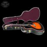An acoustic guitar with a sunburst finish displayed inside an open black hard case with a burgundy velvet interior, set against a solid black background.