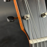 Close-up detail of an acoustic guitar headstock, showing the dark wood body, six tuning strings, silver tuning pegs, and black tuning keys against a dark gray background.