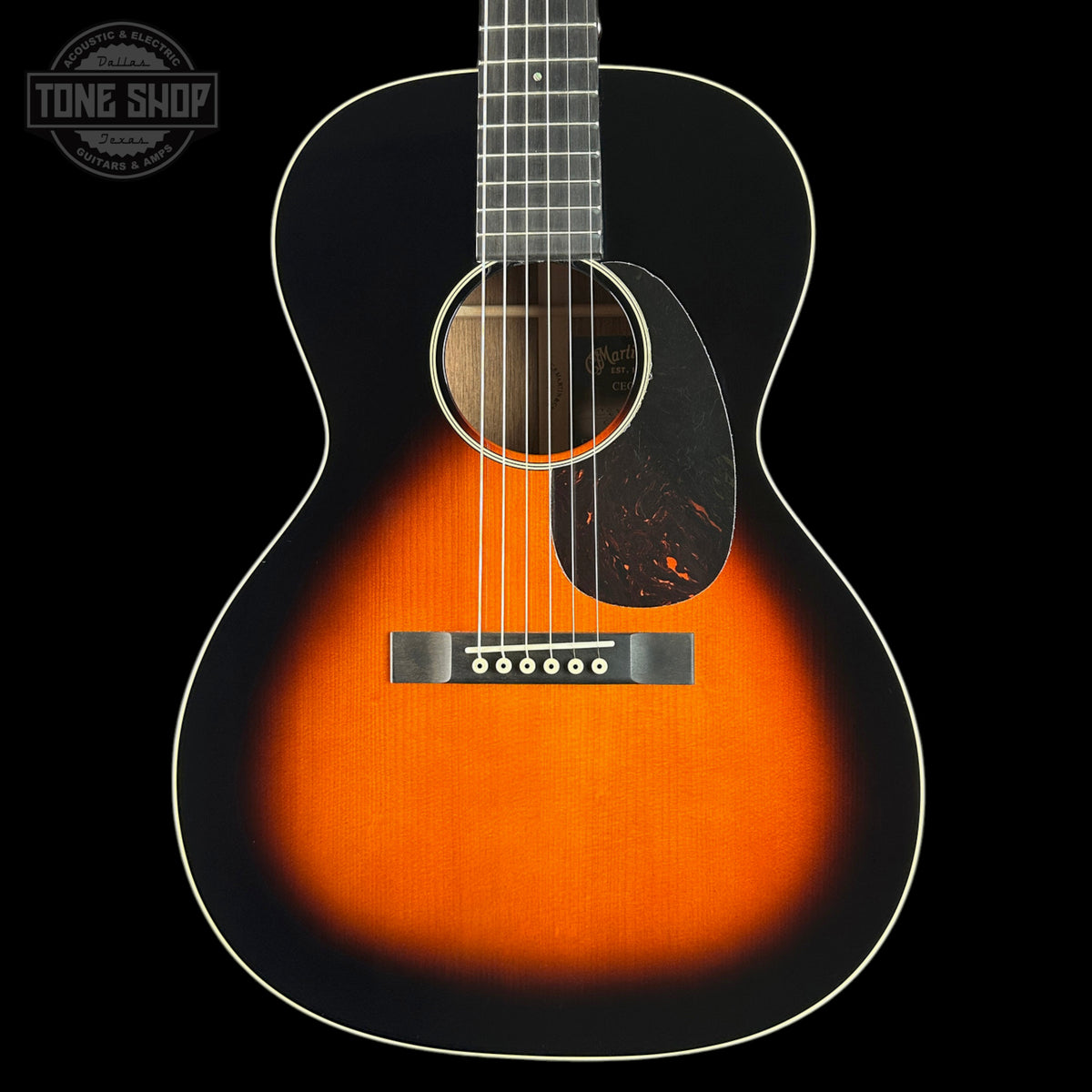 A close-up studio shot of an acoustic guitar body showing a classic sunburst finish that transitions from bright orange in the center to dark black around the edges. The guitar features white binding, a dark pickguard, and a silver bridge, set against a solid black background with a Tone Shop logo in the top-left corner.
