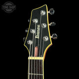 Close-up of the headstock of an Ibanez electric guitar against a black background, showing the black headstock with yellow binding, silver tuning pegs, and the white Ibanez logo.