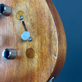 Close-up of a wooden guitar headstock corner with a small translucent white plastic cap or cover over a screw hole, set against a dark blue background.