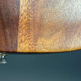 Close-up of a wooden product surface showing a warm brown wood grain finish with a small metallic knob at the bottom left against a dark blue-gray background.