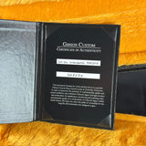 A Gibson Custom Certificate of Authenticity insert shown inside an open black leather case, placed on a textured orange fabric background. The certificate includes handwritten identification labels for a 'Gibson Standard Fokina' and serial number 'G02284'.