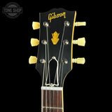 Close-up of a Gibson acoustic guitar headstock with six cream-colored tuning pegs, a gold Gibson logo, and a gold arrow emblem on a black background.