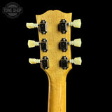 Close-up of the headstock of an acoustic guitar with a natural wood finish, featuring six black tuning pegs with cream-colored plastic knobs, set against a solid black background.