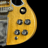 Close-up of a guitar body showing a natural wood finish, chrome control knobs, a black pickguard, and part of the tremolo system against a black background.