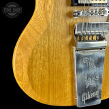 Close-up of a Gibson electric guitar body in a natural wood finish, showing the wood grain, bridge, and Gibson logo on the tailpiece against a black background.