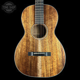 An acoustic guitar with a rich, warm brown wood grain finish, shown from the front against a solid black background. A Tone Shop watermark is visible in the top-left corner.