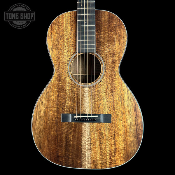 An acoustic guitar with a rich, warm brown wood grain finish, shown from the front against a solid black background. A Tone Shop watermark is visible in the top-left corner.