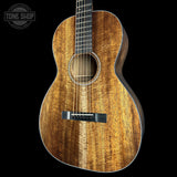A close-up studio shot of an acoustic guitar body with a rich, figured wood grain finish in warm brown and golden tones, set against a solid black background with a Tone Shop watermark in the top left corner.