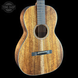 An acoustic guitar with a rich, warm brown wood grain finish, shown from a front three-quarter angle against a solid black background. A Tone Shop watermark is visible in the top-left corner.