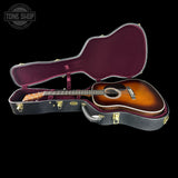 An acoustic guitar with a sunburst finish displayed inside an open black hard case with a burgundy interior, set against a solid black background. A 'TONE SHOP' watermark is visible in the top-left corner.