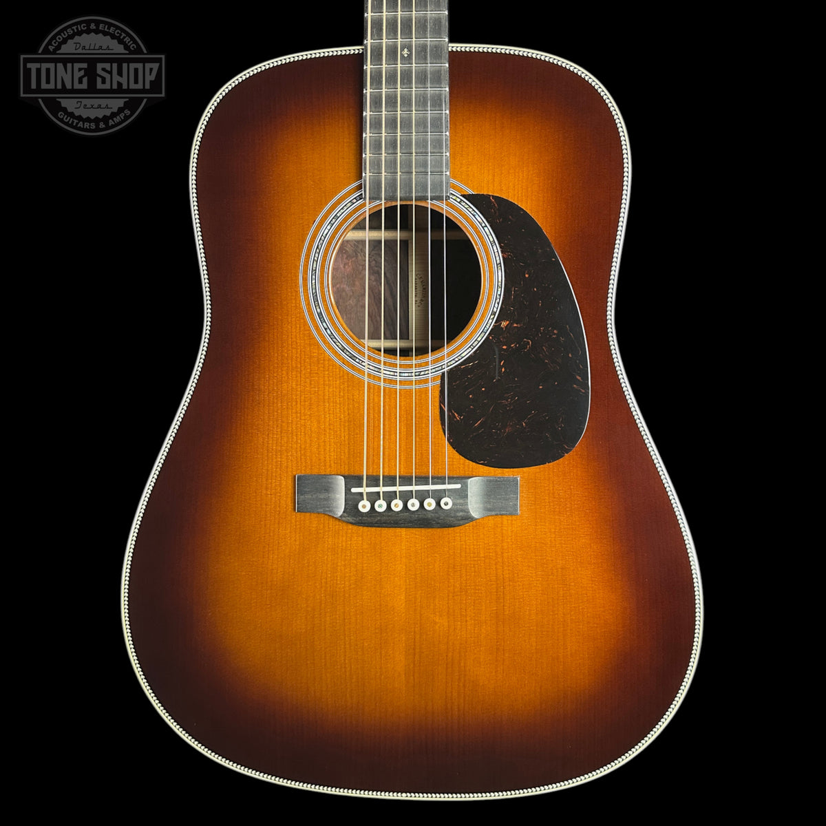 A classic acoustic guitar with a sunburst finish, featuring a warm orange and brown gradient, a black pickguard, and white binding, shown against a solid black background with a Tone Shop logo in the top left corner.