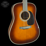 A close-up of a wooden acoustic guitar body with a sunburst finish, showing the sound hole, pickguard, bridge, and part of the neck against a black background. A Tone Shop logo watermark is visible in the top-left corner.
