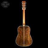 The back of an acoustic guitar with a rich, figured wood backplate and a matching wood neck, shown against a solid black background with a Tone Shop watermark in the top left corner.